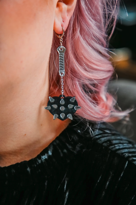 A mace weapon acrylic earring with a black love heart shaped ball and silver handle and spikes against pink hair