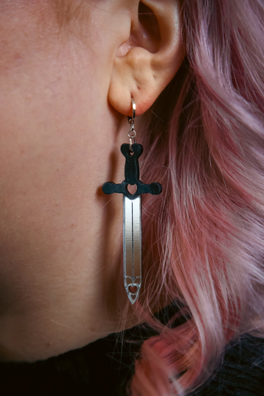 A sword acrylic earring with a black handle and reflective silver blade against pink hair