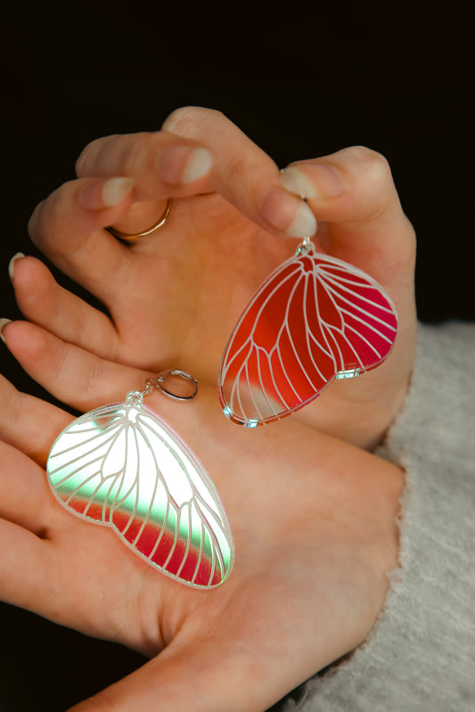 A pair of iridescent acrylic earring styled after a fairies wing being held by hands
