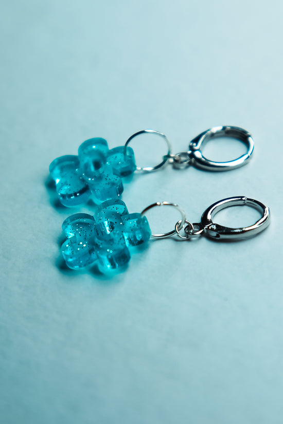 A pair of transparent glitter blue flower acrylic earring against a blue background