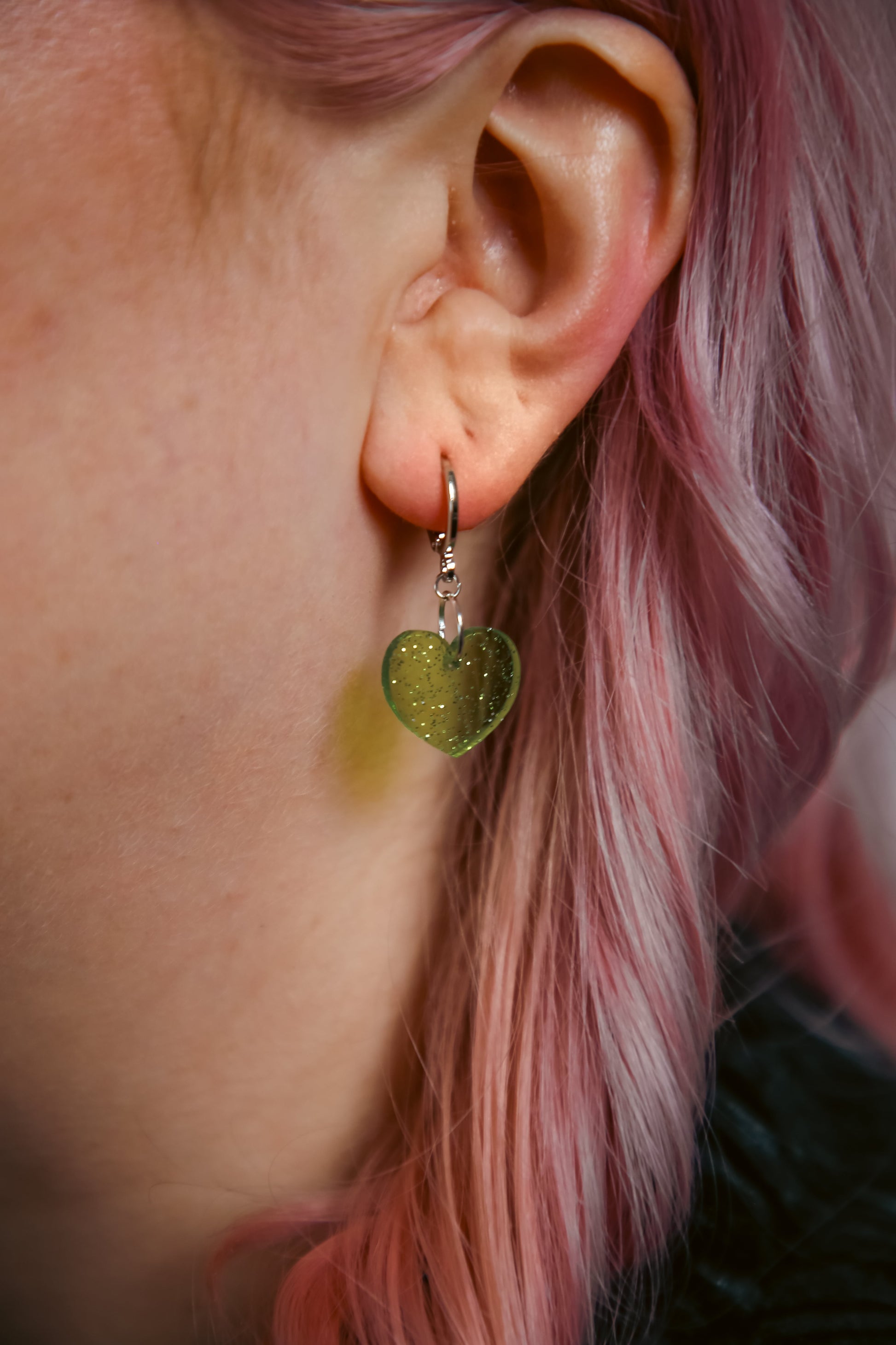 A transparent glitter green heart acrylic earring against a model with pink hair
