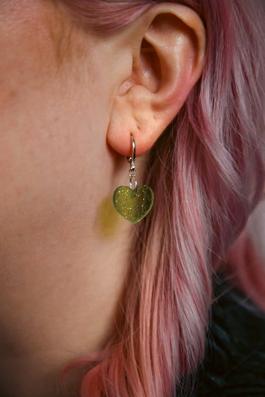 A transparent glitter green heart acrylic earring against a model with pink hair