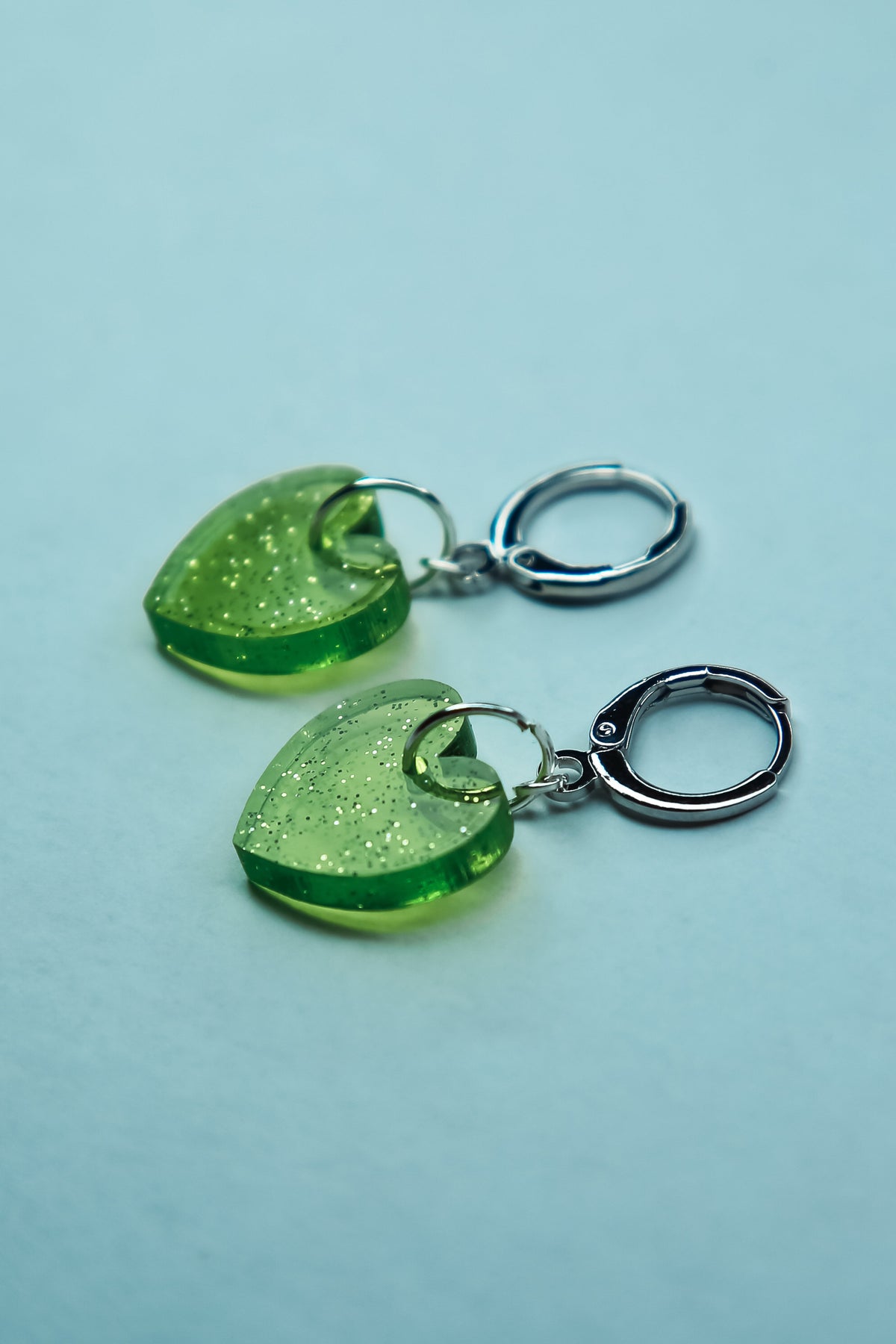 A pair of transparent glitter green heart acrylic earring against a blue background