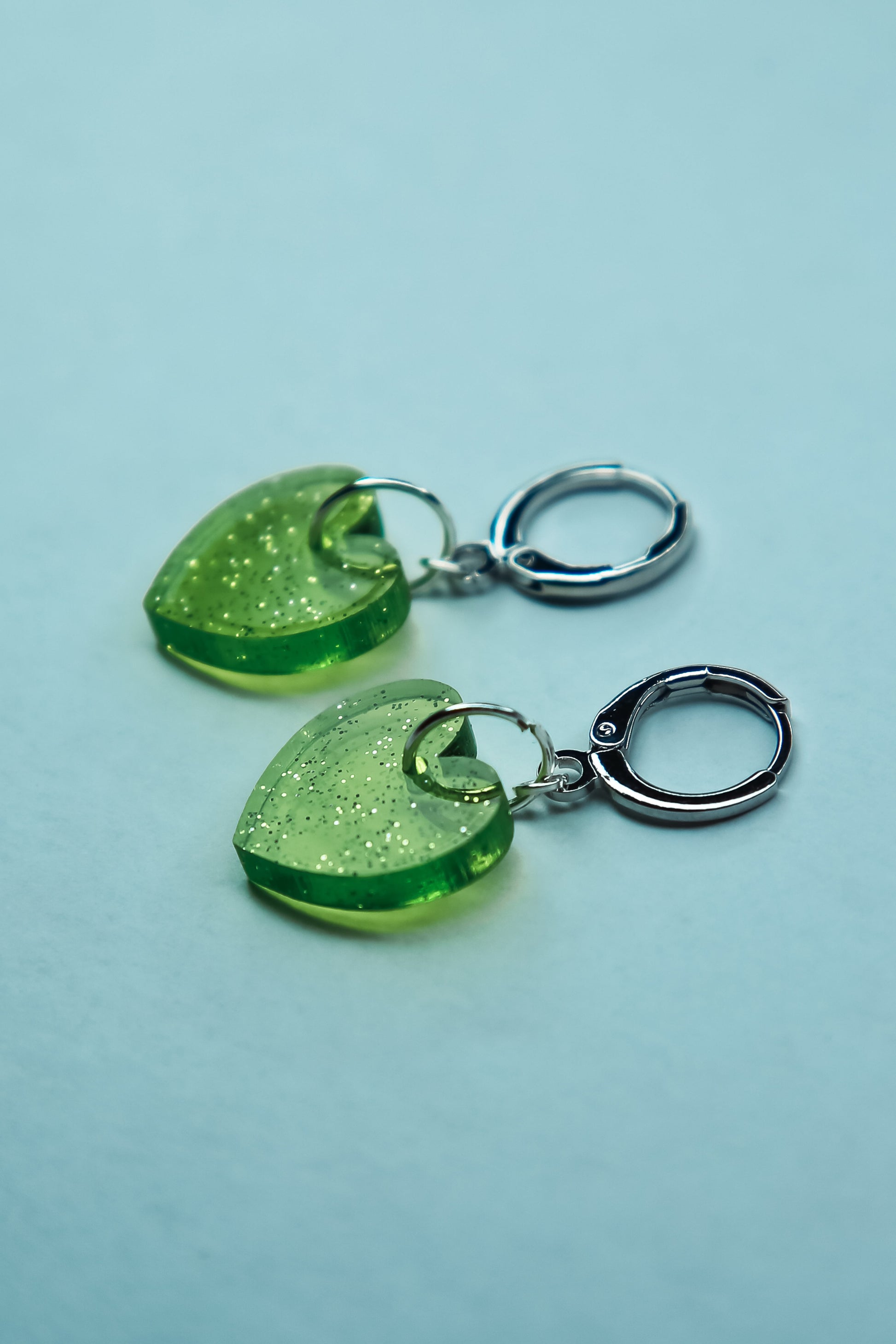 A pair of transparent glitter green heart acrylic earring against a blue background