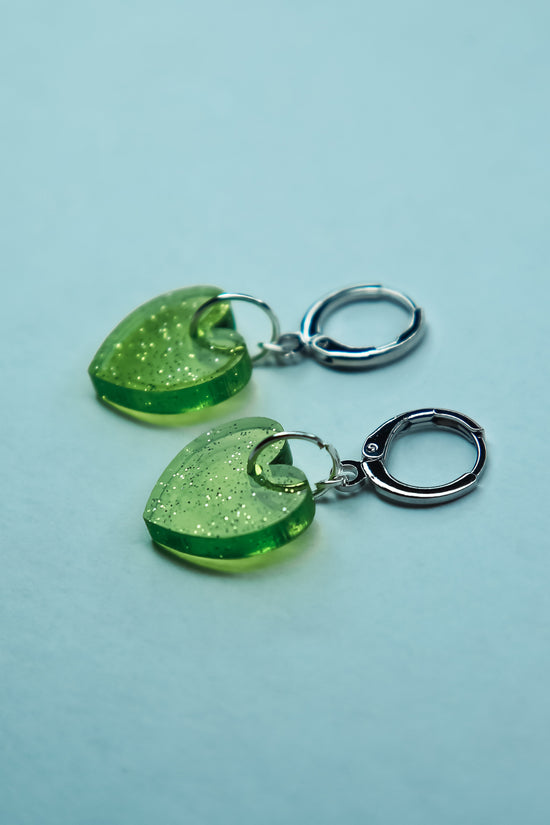 A pair of transparent glitter green heart acrylic earring against a blue background