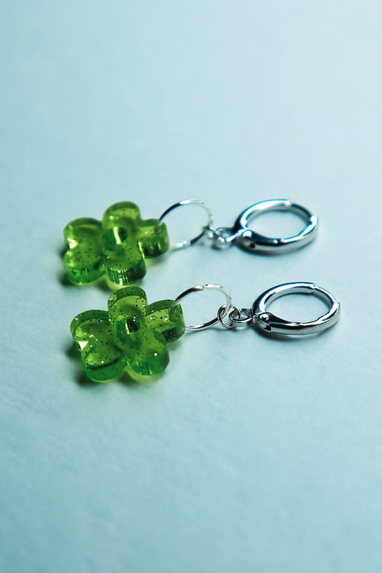A pair of transparent glitter green flower acrylic earring against a blue background
