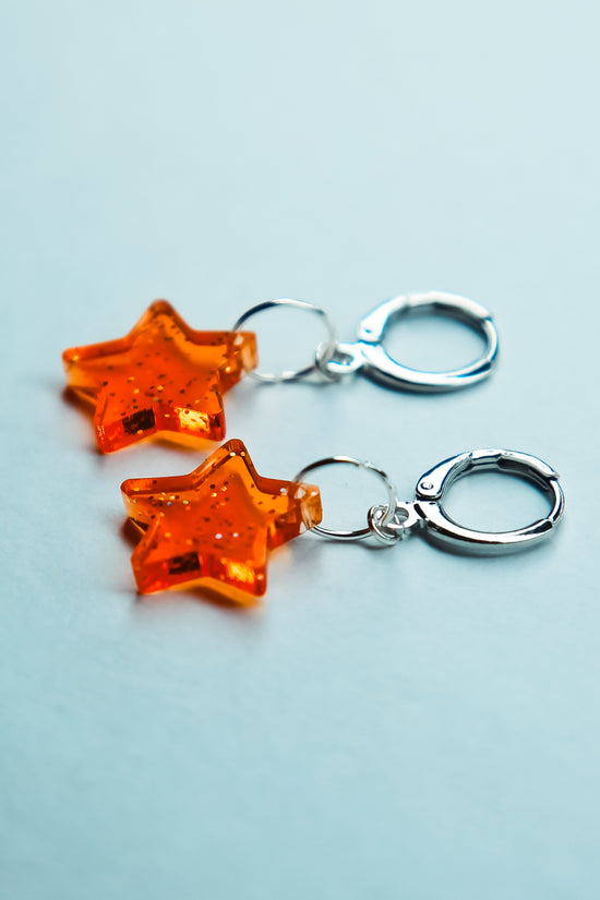 A pair of transparent glitter orange star acrylic earrings on a blue background