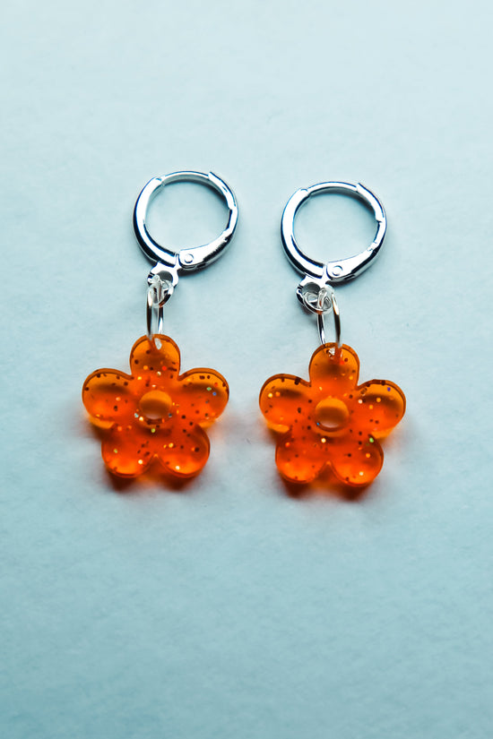 A pair of transparent glitter orange flower acrylic earrings on an ear