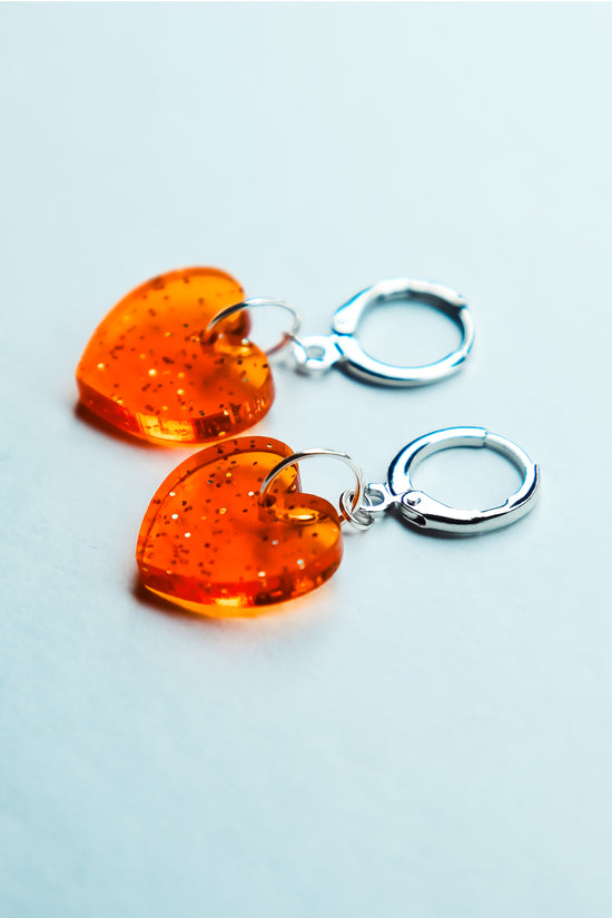 A pair of transparent glitter orange heart acrylic earrings on a blue background