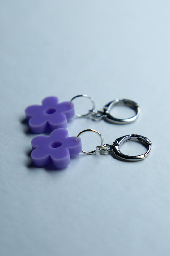 A pair of pastel purple flower acrylic earring on a blue background