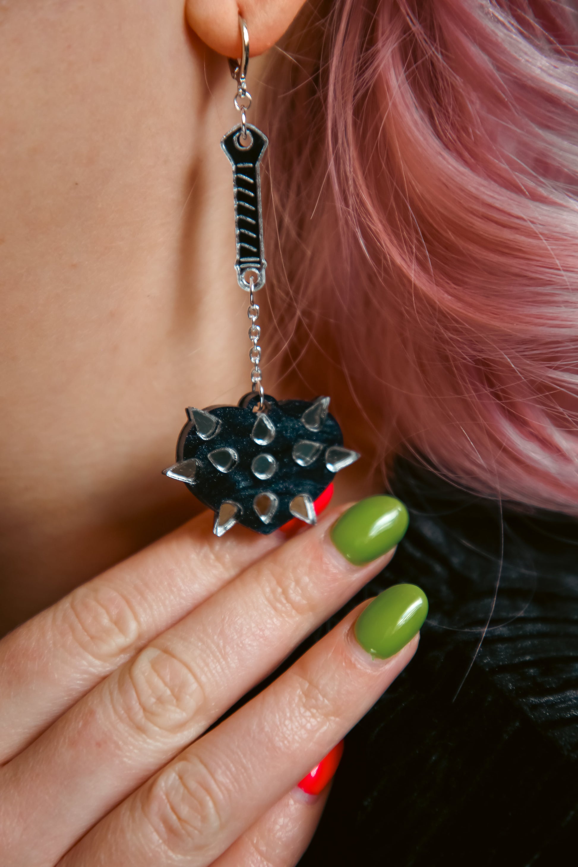 A mace weapon acrylic earring with a black love heart shaped ball and silver handle and spikes against pink hair propped up by a hand with red and green nails