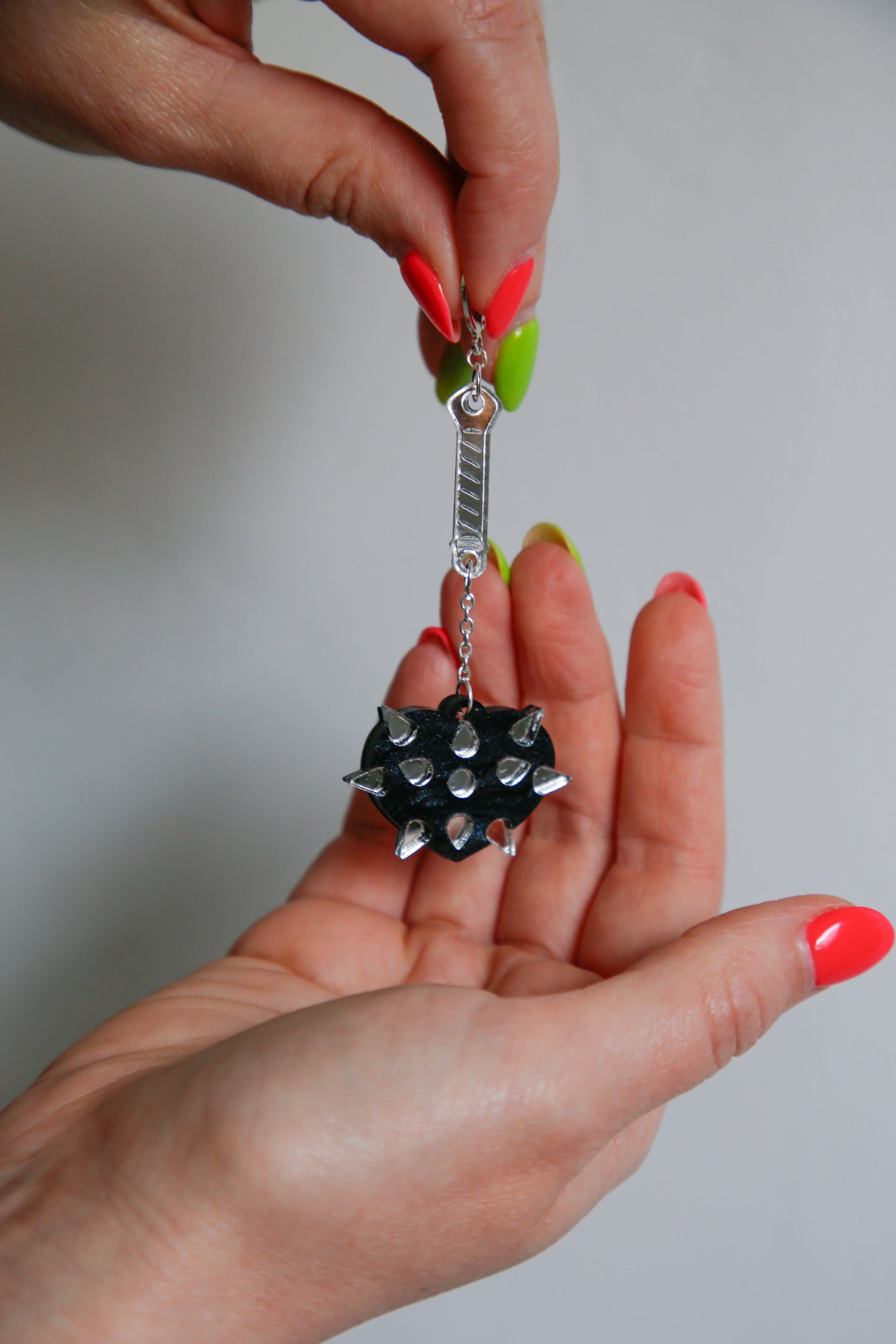 A mace weapon acrylic earring with a black love heart shaped ball and silver handle being held up