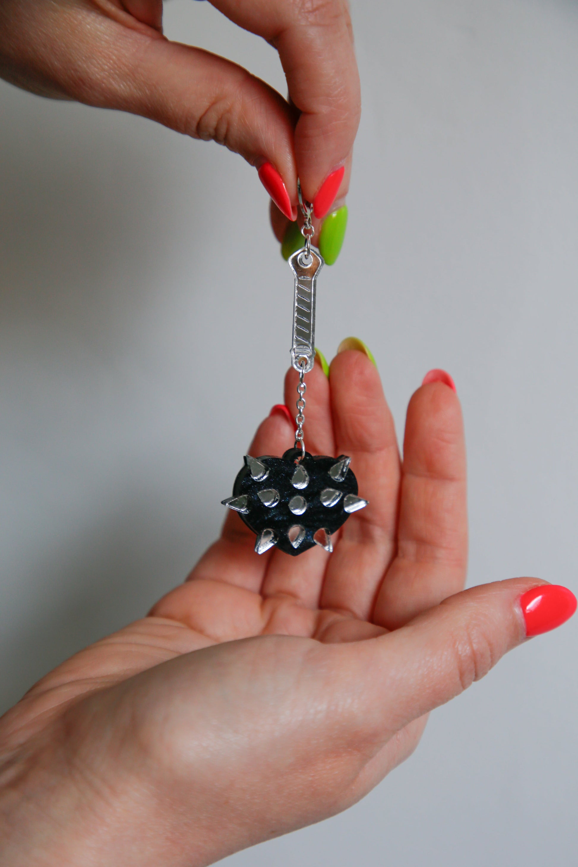 A mace weapon acrylic earring with a black love heart shaped ball and silver handle being held up