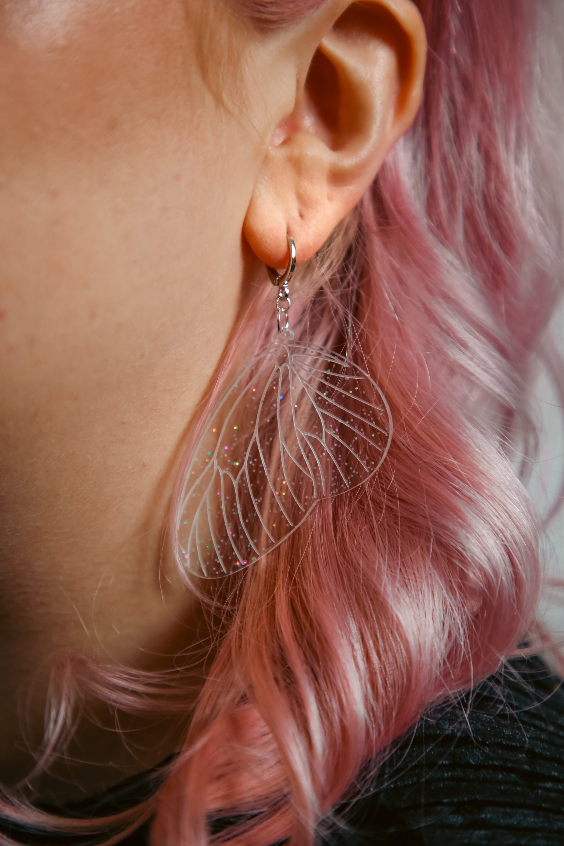 glitter transparent fairy wing earring on a persons ear with pink hair behind
