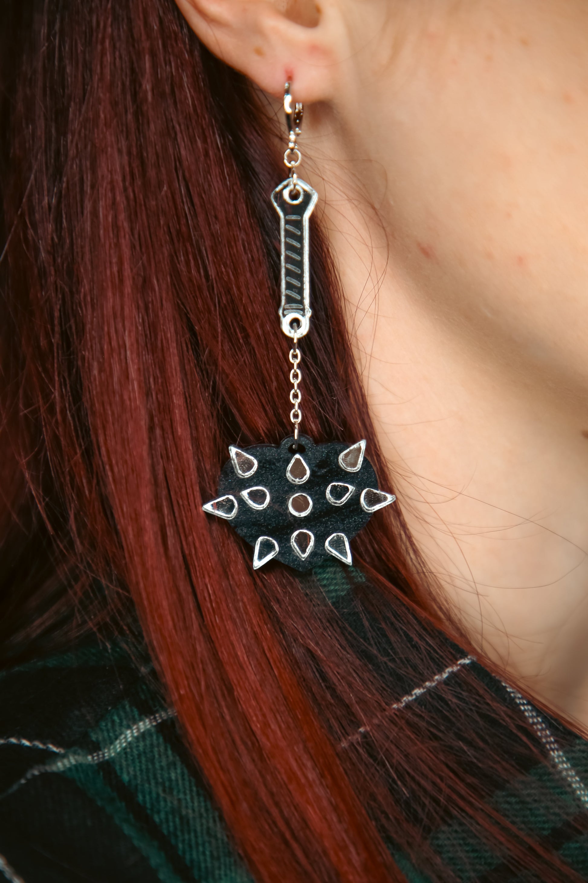 A mace weapon acrylic earring with a black love heart shaped ball and silver handle and spikes against brown hair