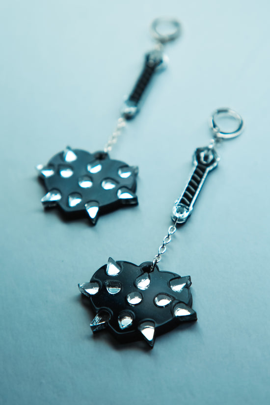 A flat lay of a pair of mace weapon acrylic earring with a black love heart shaped ball and silver handle and spikes