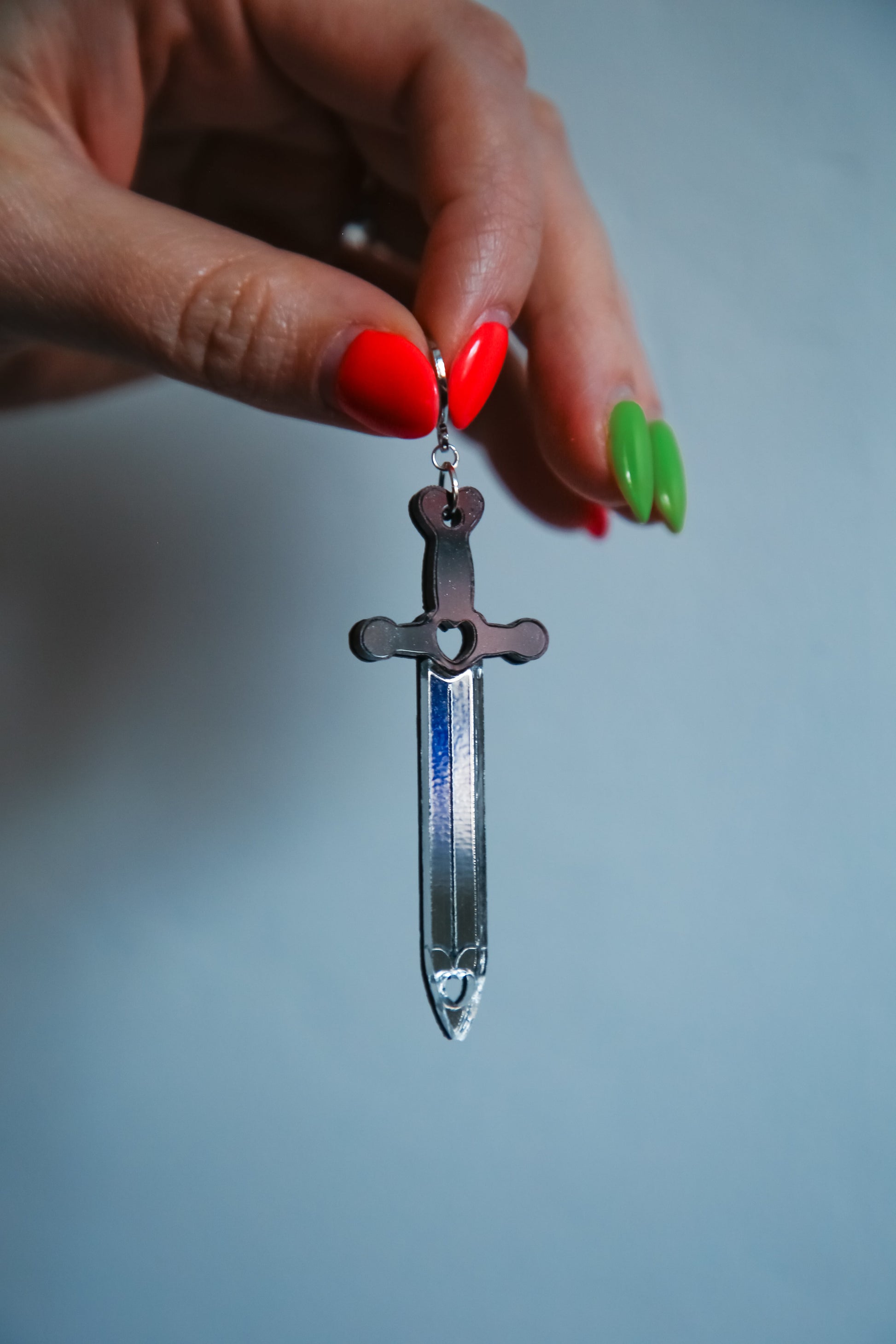 A sword acrylic earring with a black handle and reflective silver blade being held up by a hand