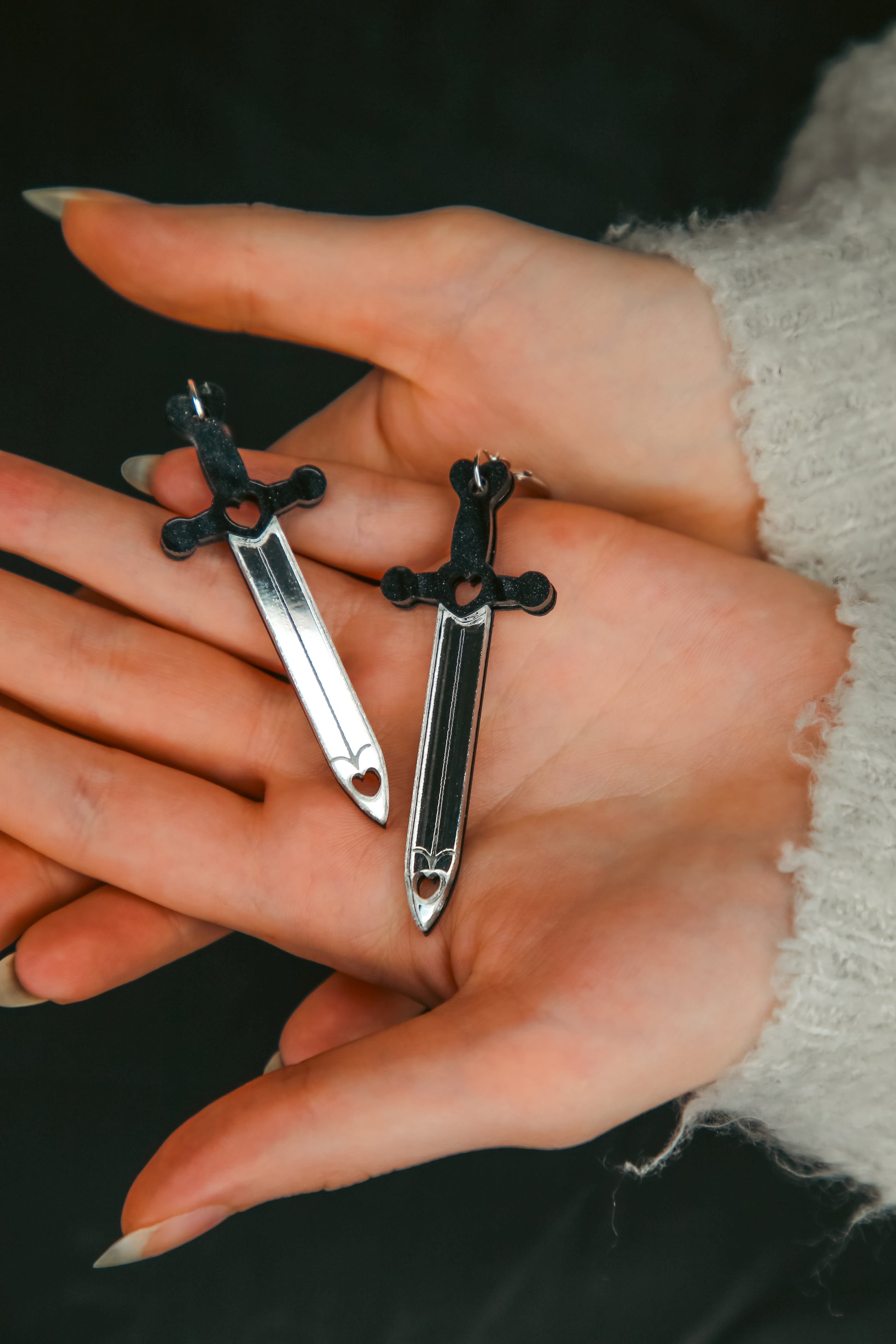 A pair of sword acrylic earrings with a black handle and reflective silver blade being held by a hand