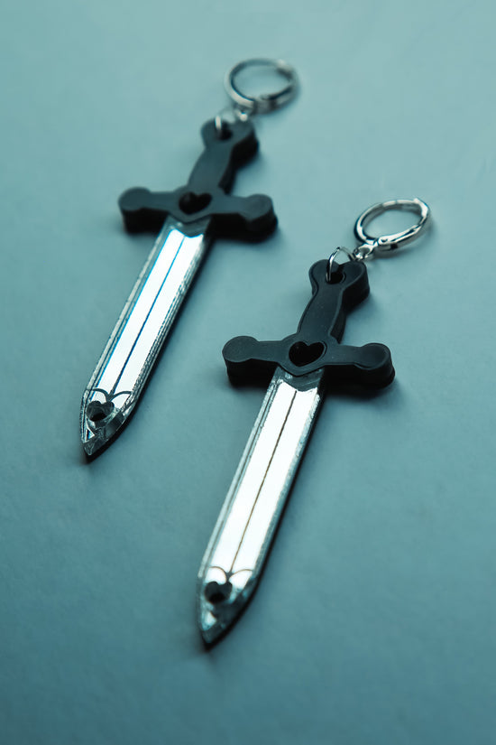 A flat lay of a pair of sword acrylic earring with a black handle and reflective silver blade 