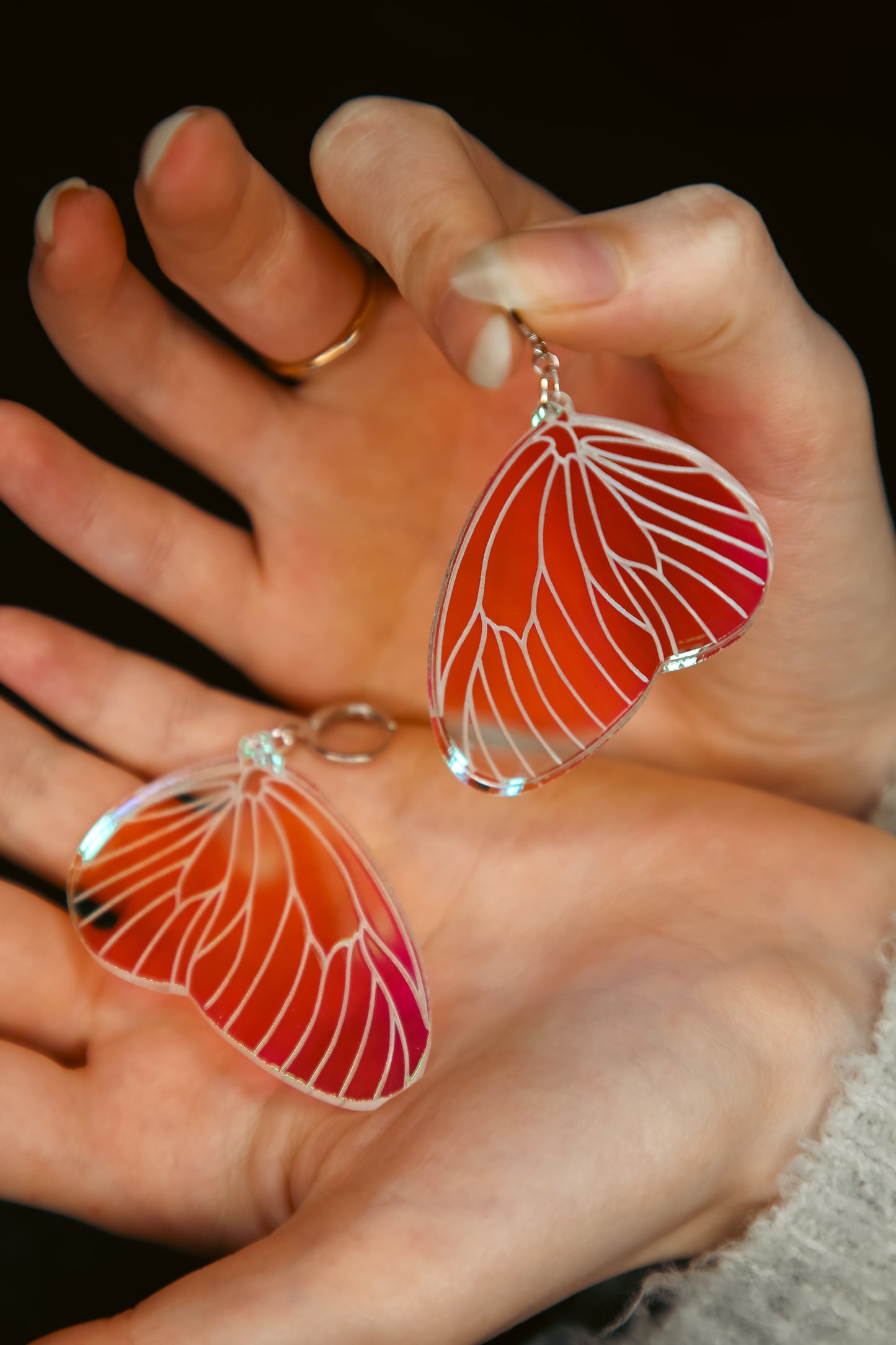 A pair of iridescent acrylic earring styled after a fairies wing being held by hands