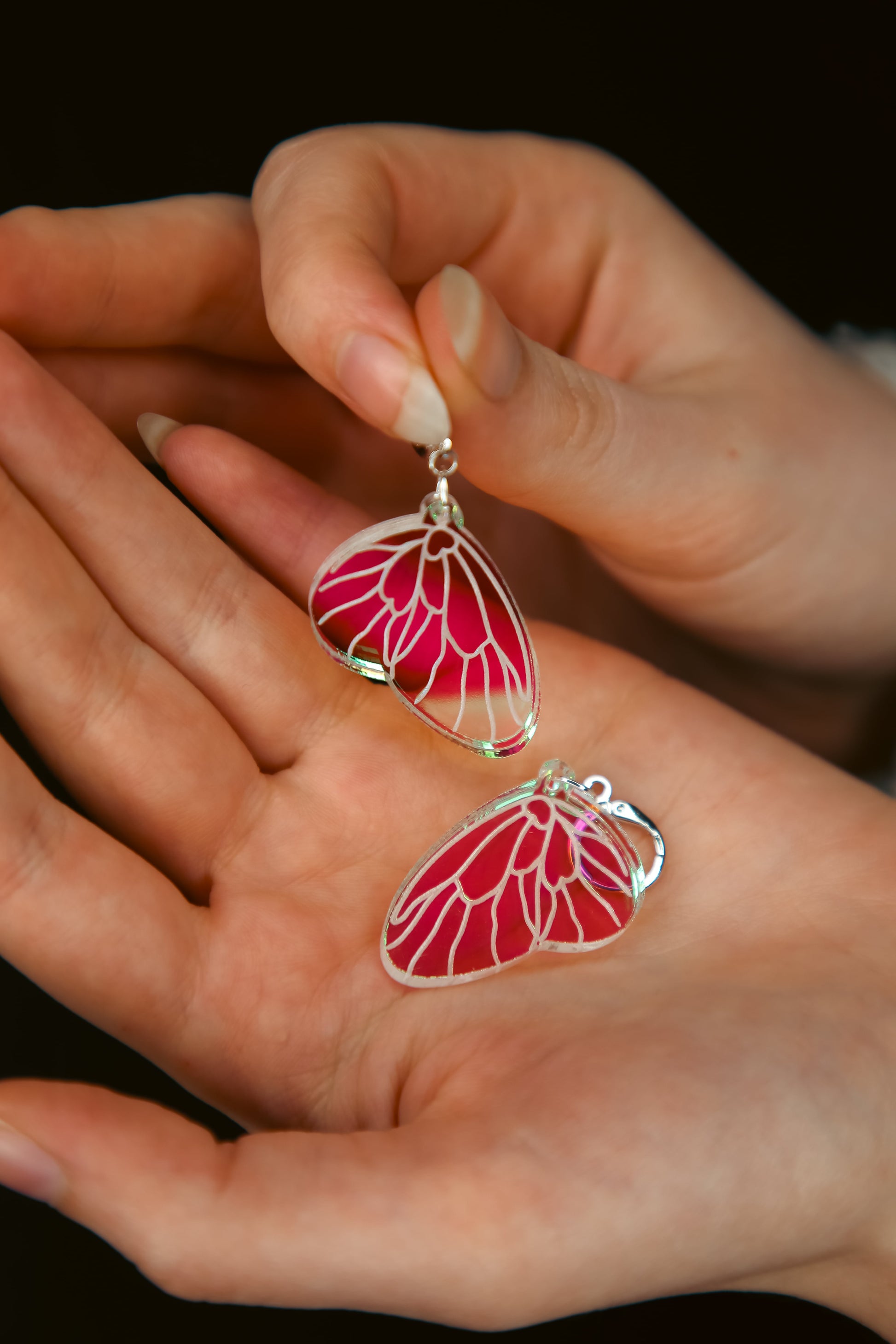 A pair of mini iridescent acrylic earring styled after a fairies wing being held by hands