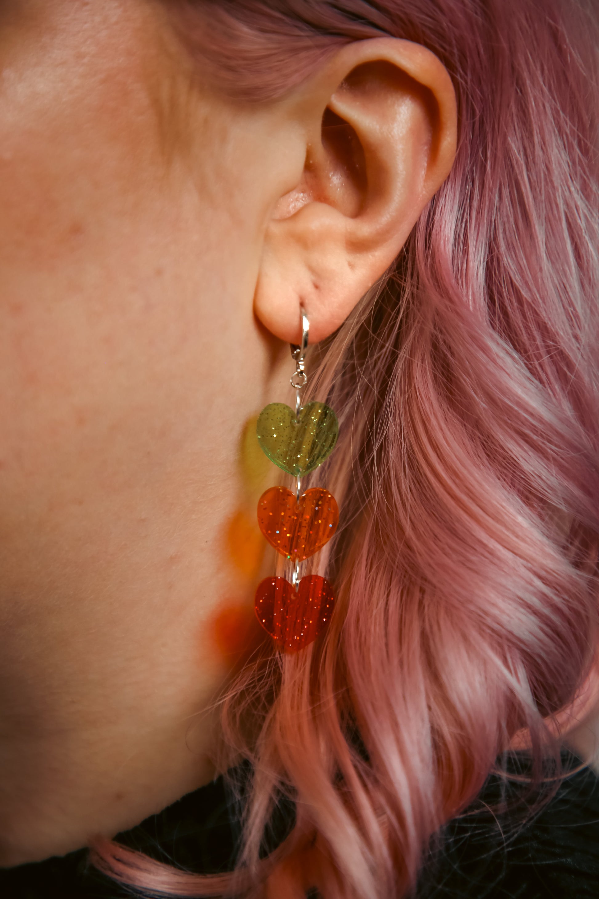 Acrylic earring which is a stack of transparent and glittery green, orange and red hearts against pink hair