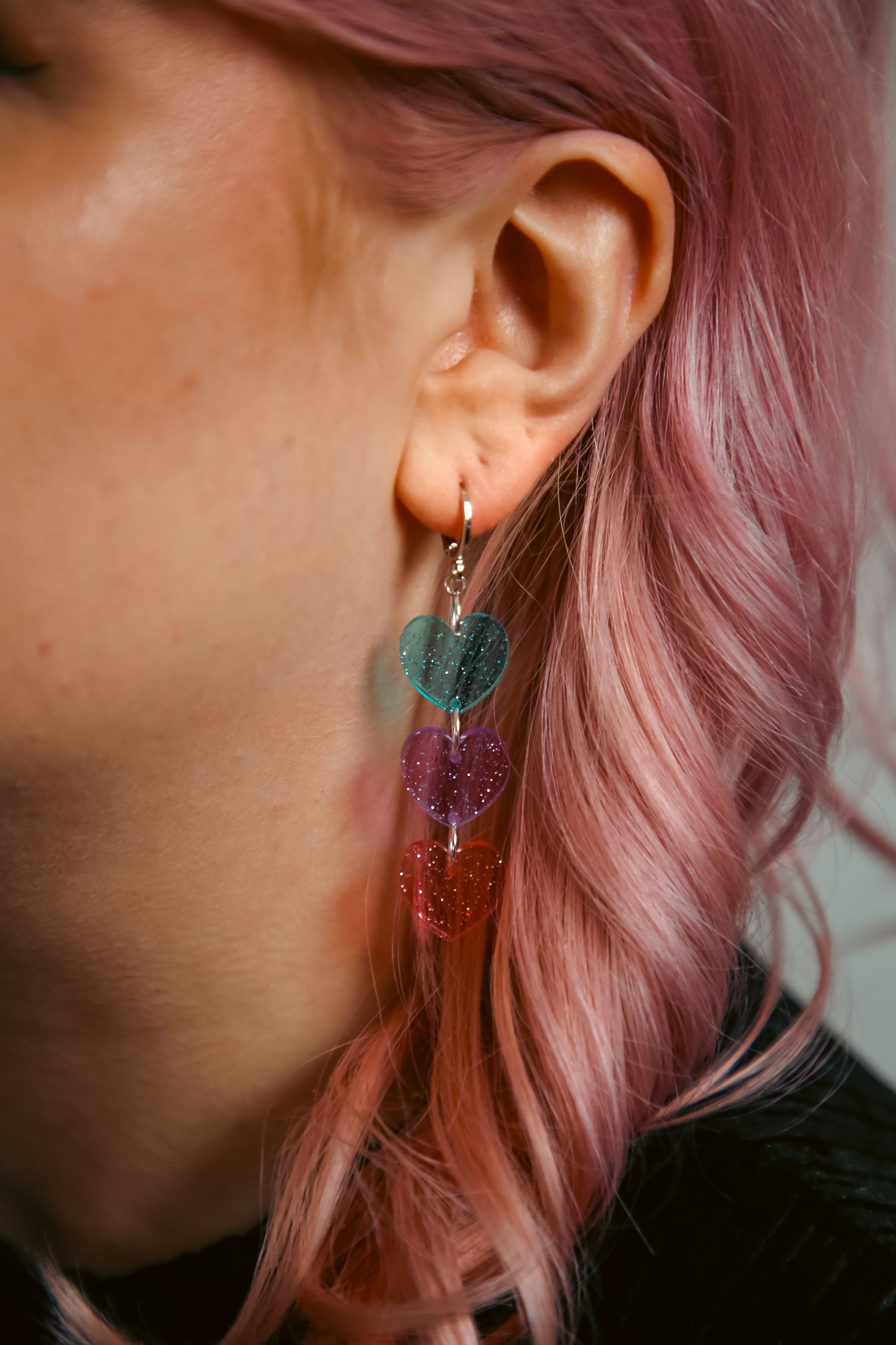 Acrylic earring which is a stack of transparent and glittery blue, pruple and pink hearts against pink hair