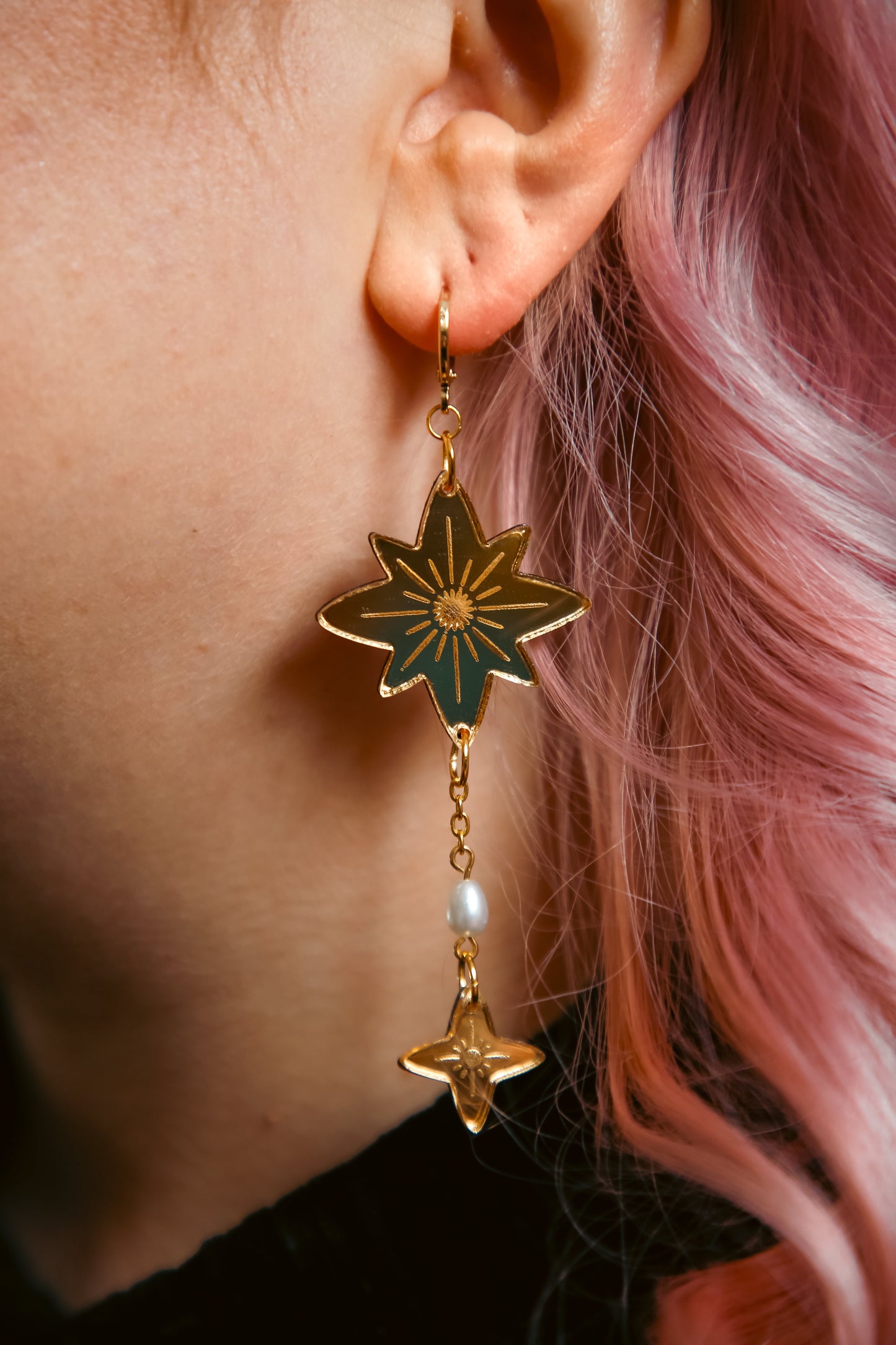 big and small star shaped acrylic earring made from reflective gold acrylic with a pearl dangling off against pink hair