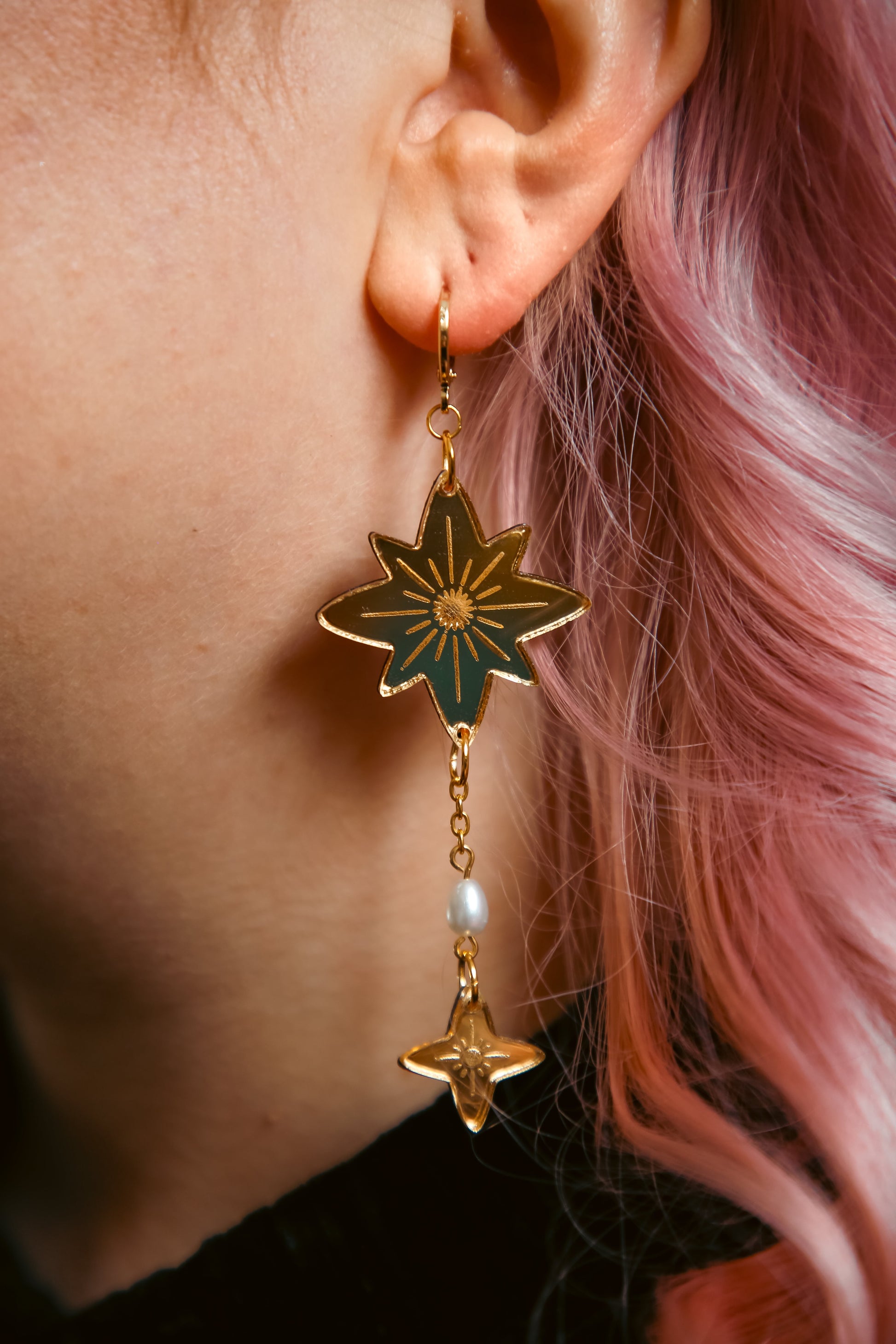 big and small star shaped acrylic earring made from reflective gold acrylic with a pearl dangling off against pink hair