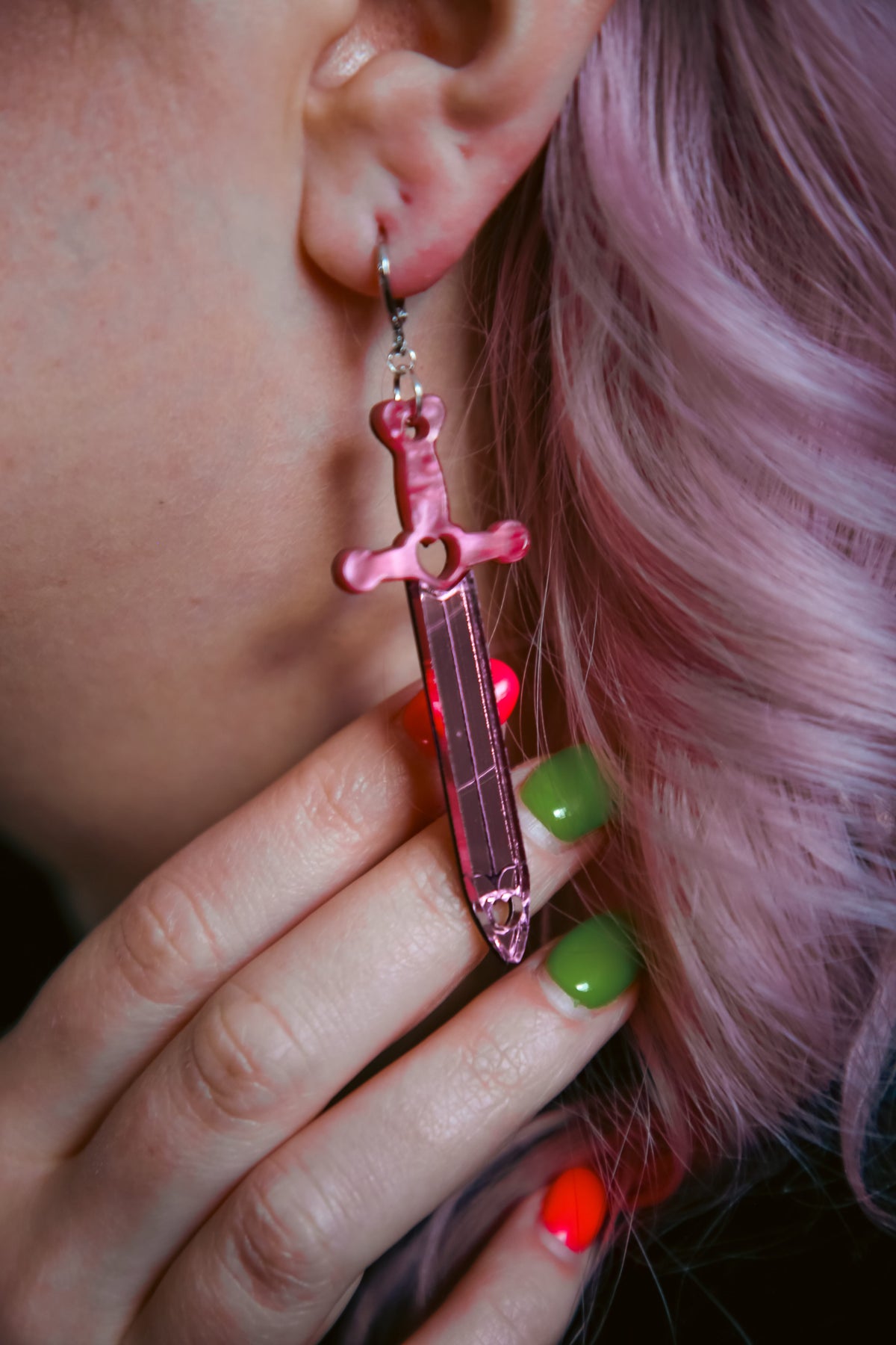 Pink sword acrylic earring with a reflective pink blade against pink hair and red and green finger nails