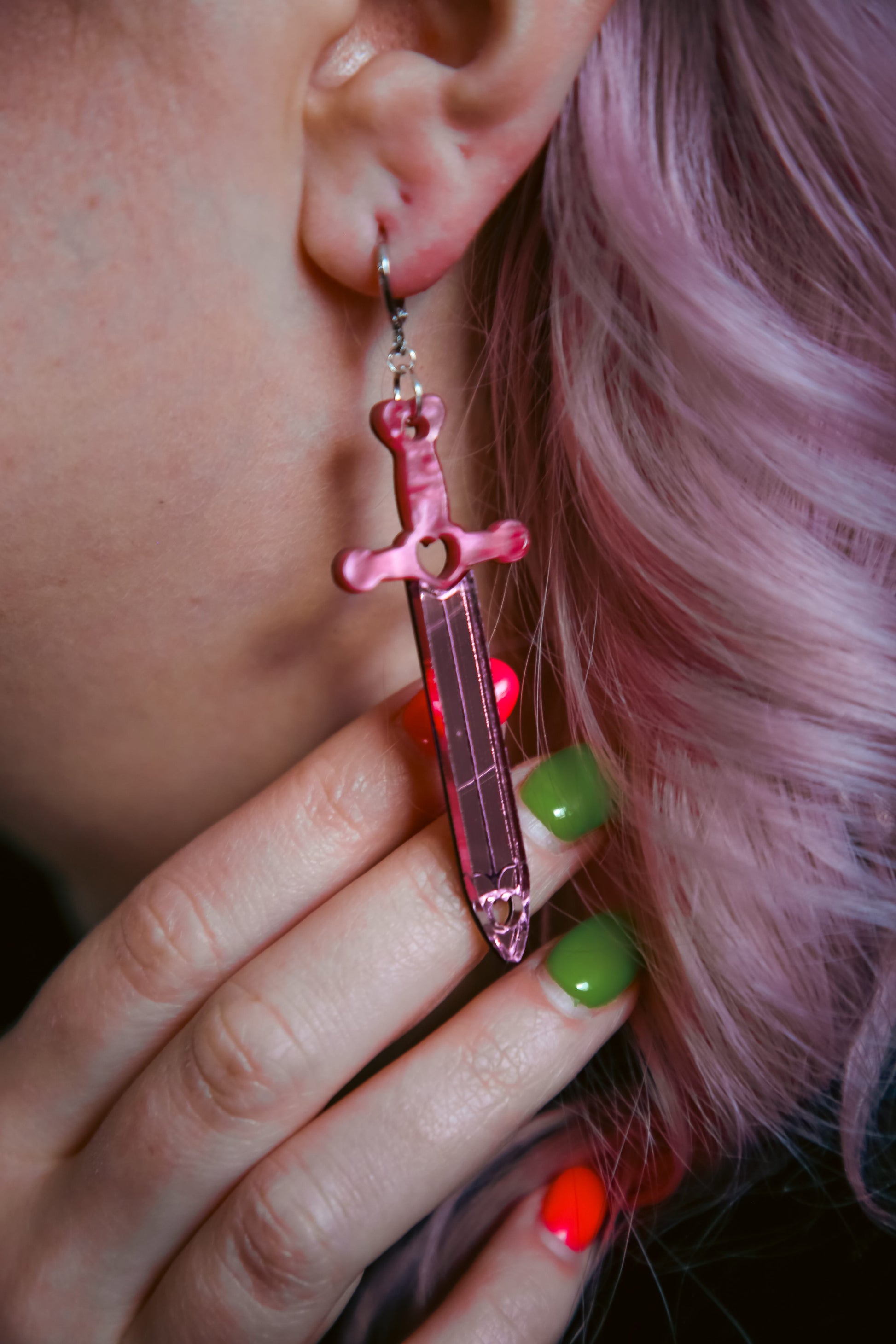 Pink sword acrylic earring with a reflective pink blade against pink hair and red and green finger nails