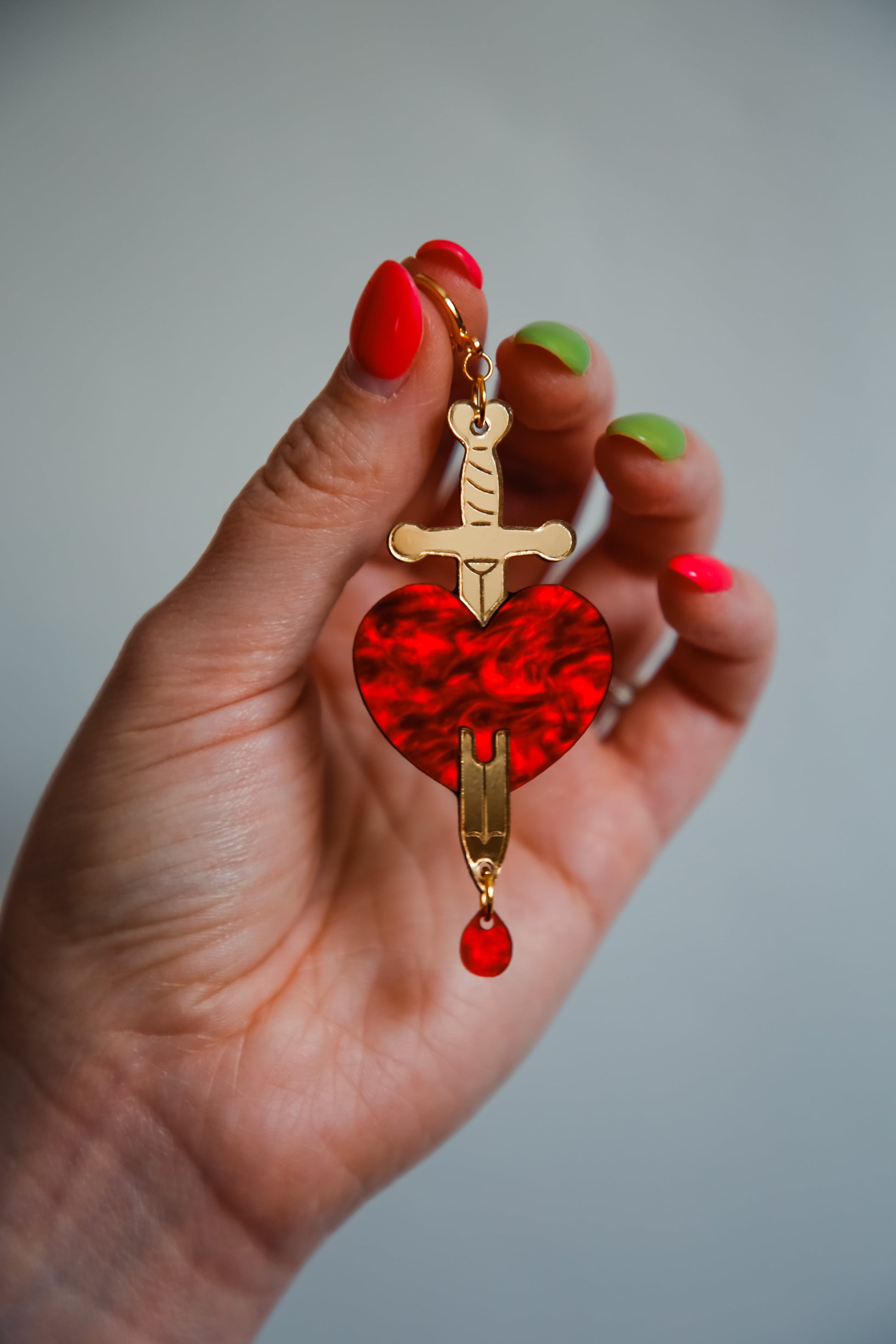 red and gold sword and heart acrylic earring being held by a hand