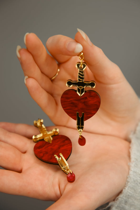 a pair of red and gold sword and heart acrylic earring being held by hands