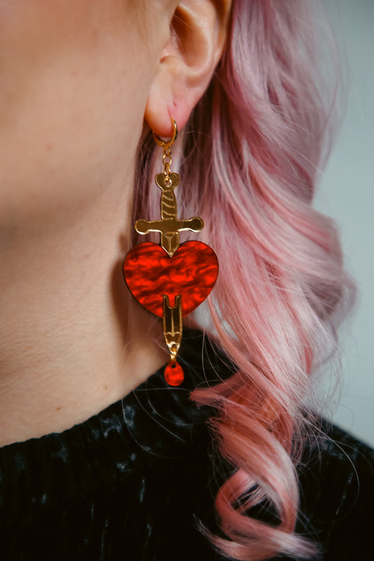 red and gold sword and heart acrylic earring being worn by a person with pink hair