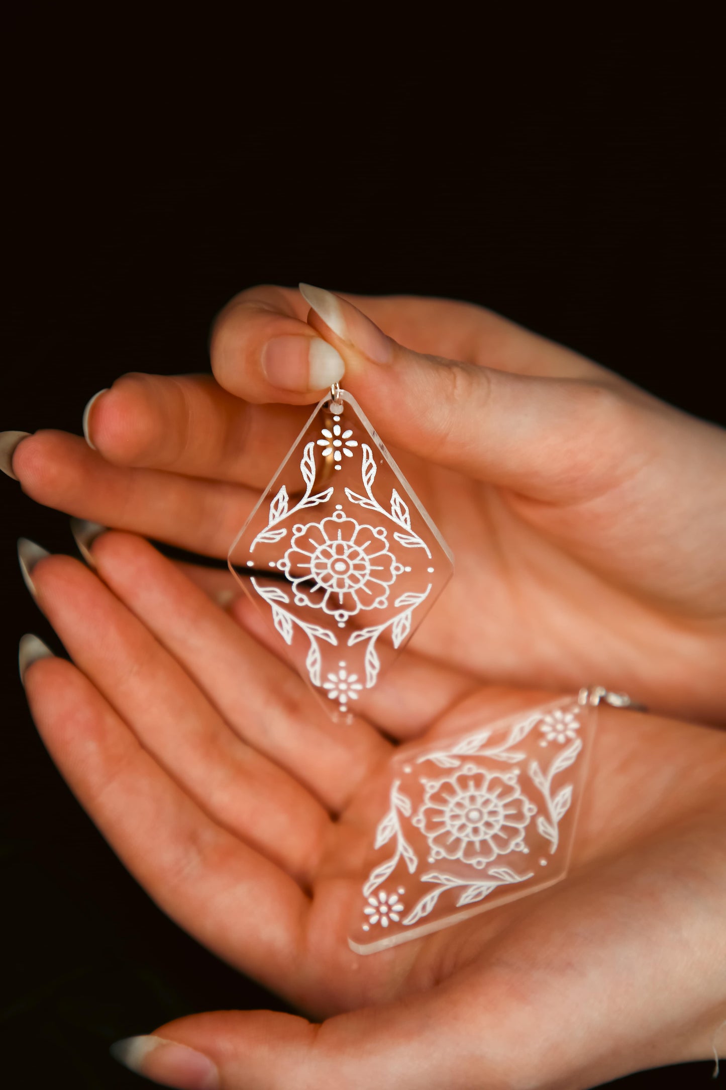 a pair of flower inspired acrylic earring made from transparent acrylic with a white design being held by hands